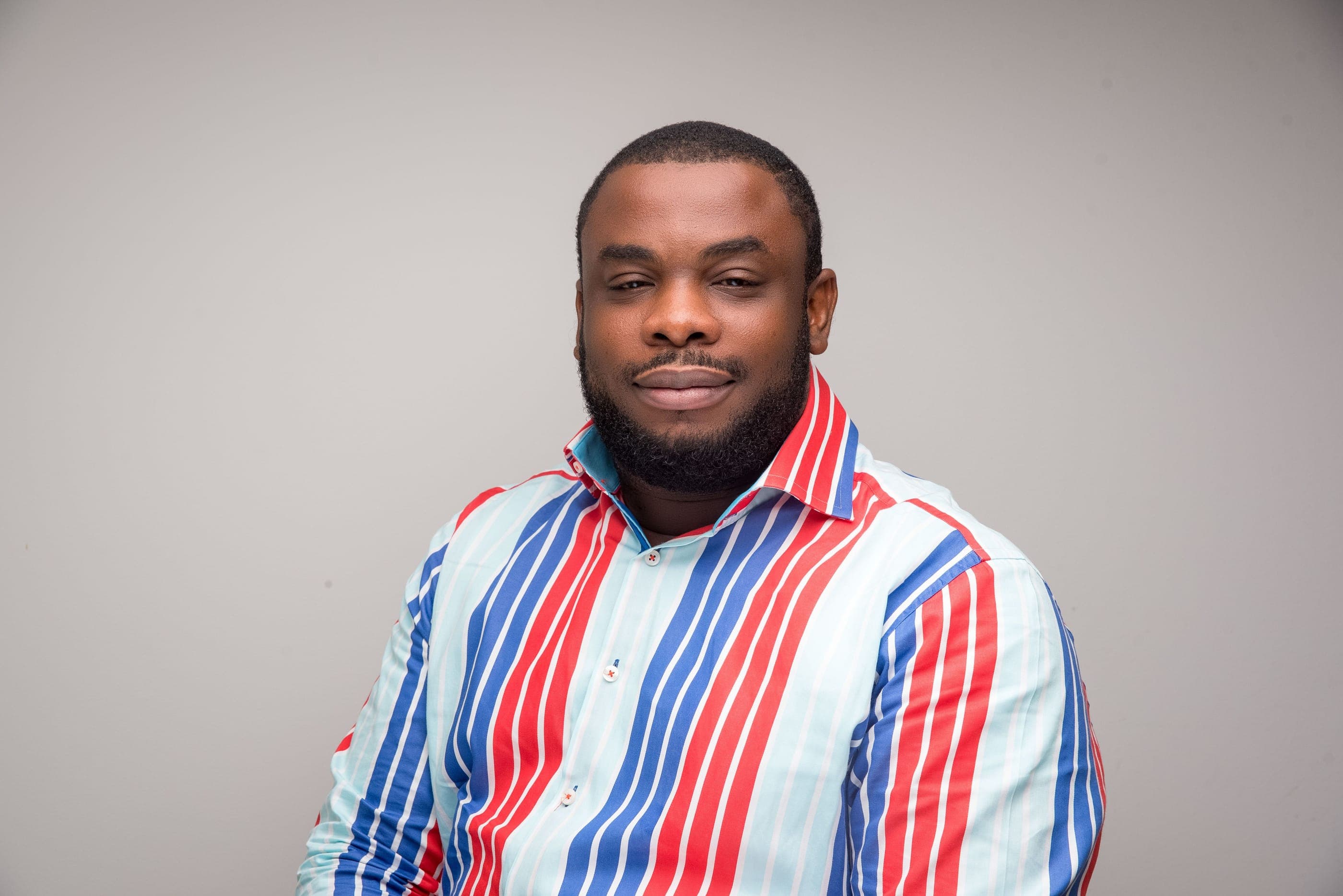 Olayiwola Allen, smiling, in a red, white and blue striped shirt against a light grey backdrop